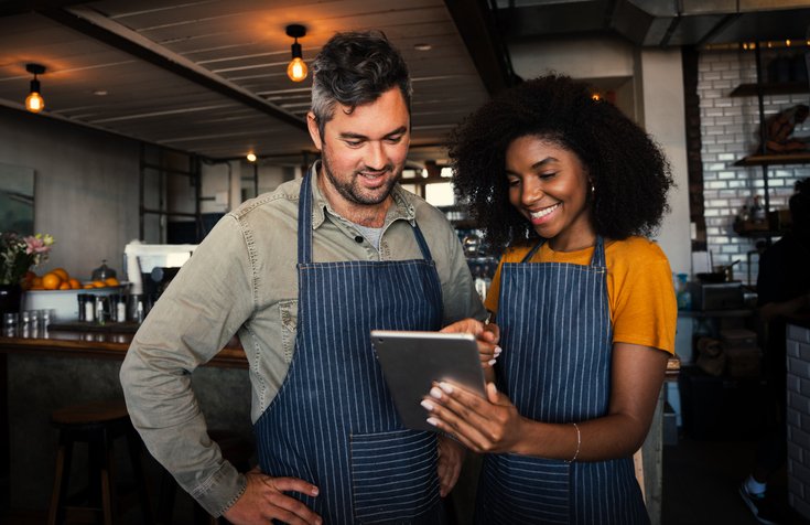 Male and female coffee shop owners smiling at tablet in coffee shop